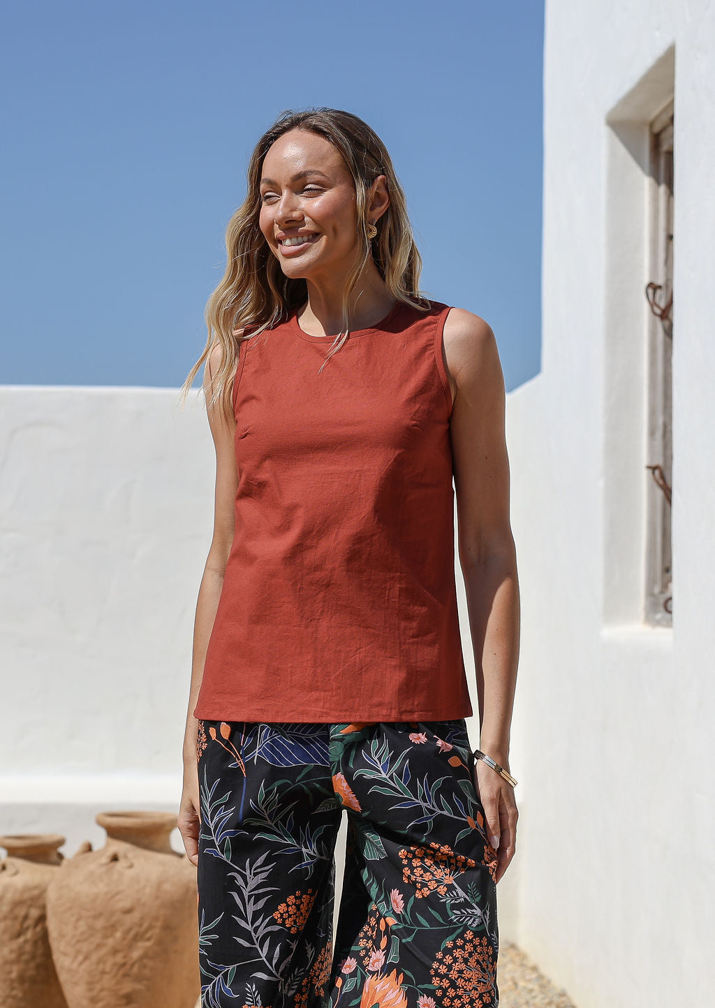 Woman wearing a rust colored sleeveless 100% cotton top and dark floral pants standing against a white wall.