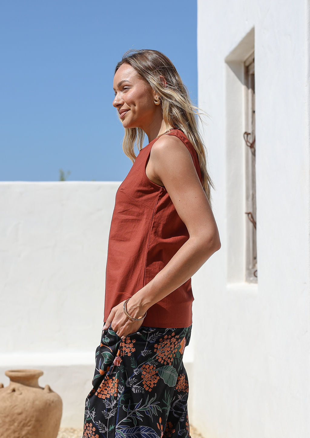 Woman wearing a relaxed fit rust-colored sleeveless cotton top with side bust darts  and floral pants standing outdoors in front of white wall