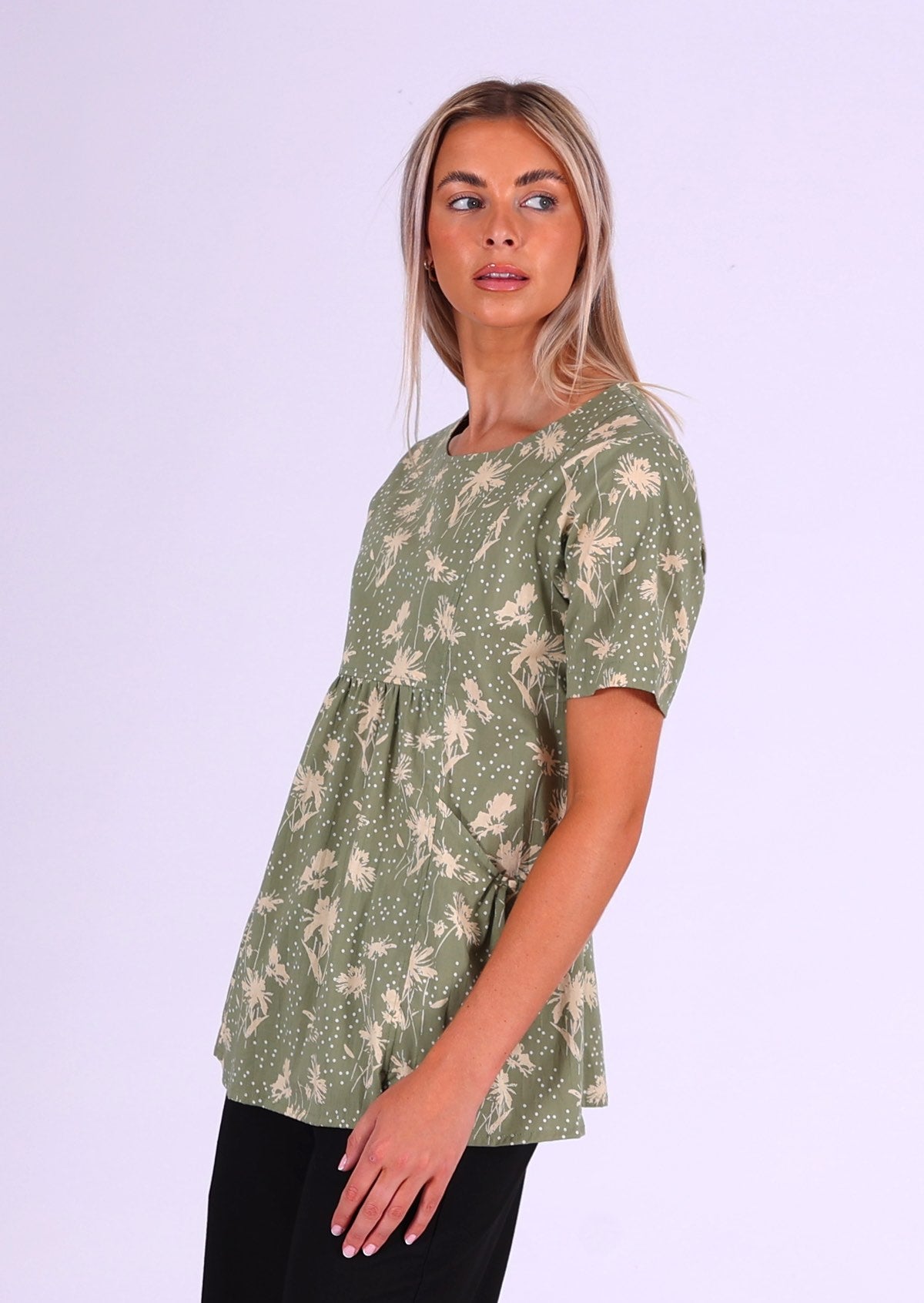 Woman stands sideways in cotton relaxed fit short sleeve top with gathers under the bust and generous pockets