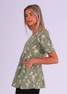 Woman stands in cotton cream on green floral print relaxed fit top with high round neckline, short sleeves and hands in generous pockets