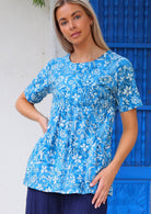 Woman stands in cotton blue floral print relaxed fit top with high round neckline, short sleeves and generous pockets