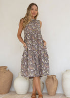 Woman in a sleeveless, floral light brown cotton dress standing in front of decorative vases with both the hands in pockets.