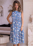 Woman poses in a sleeveless tiered cotton dress with pockets and coral pink, sage green and lemon yellow florals on dusty blue base.