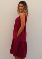 Woman stands sideways in sleeveless cotton midi length dress in deep red with hands in deep pockets