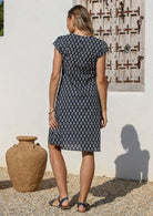 Woman standing in back pose wears a knee length navy blue floral printed cotton dress standing in front of a rustic door with clay pots nearby