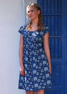 Woman with blonde hair wearing a blue cotton dress with cap sleeves in light blue and white floral design, in front of blue door
