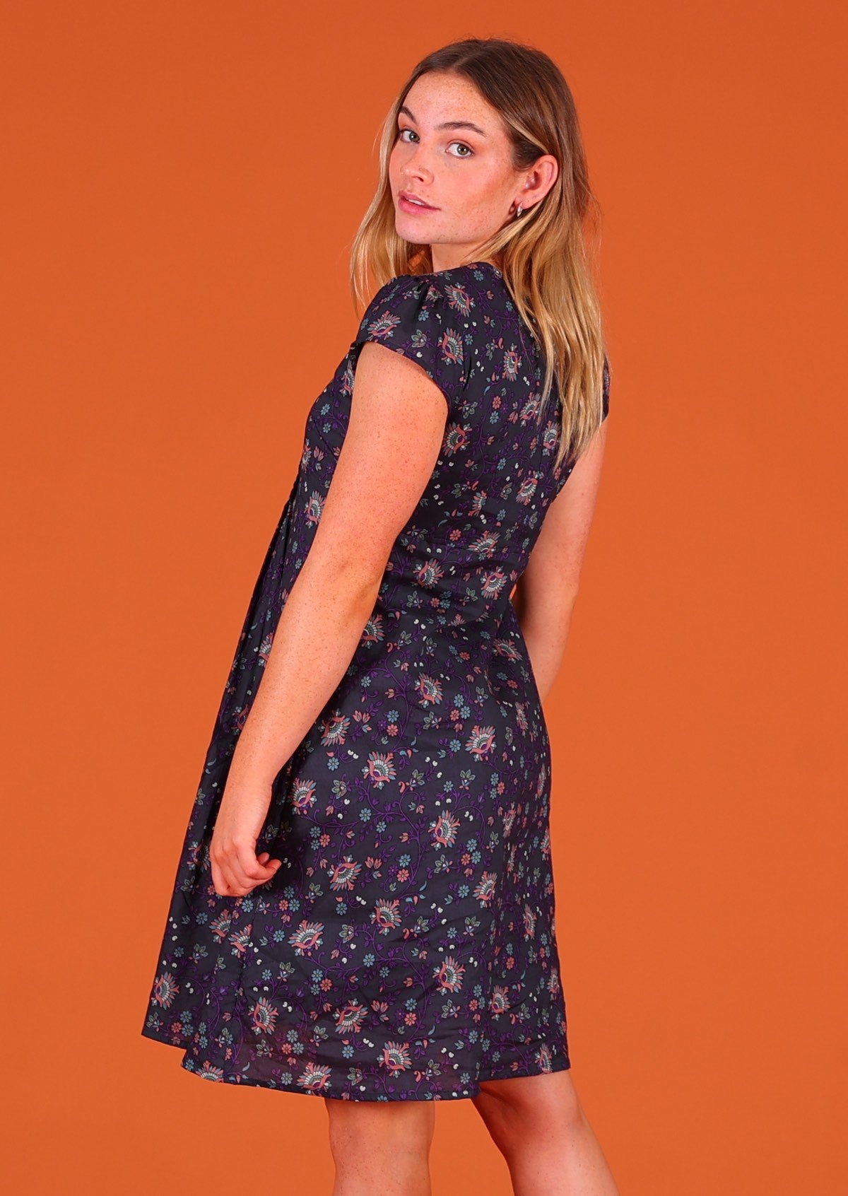 Model standing looking back over her shoulder wearing 100 percent cotton short sleeve pleated sundress in deep purple Indian floral