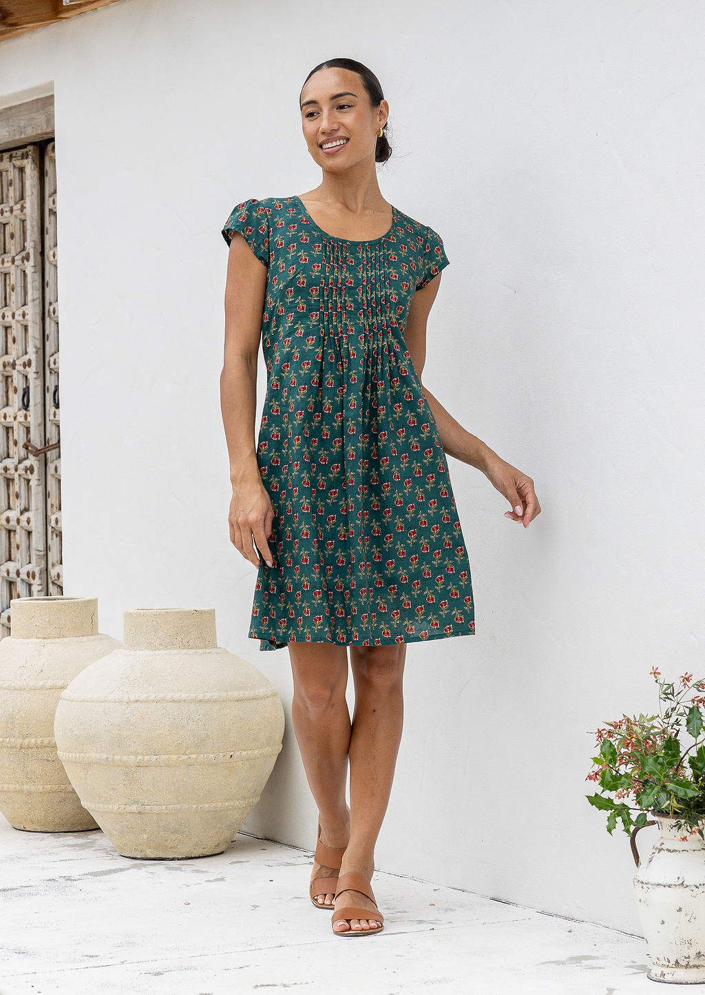 Woman wearing a green cotton patterned sundress standing in a room with white walls and decorative vases.