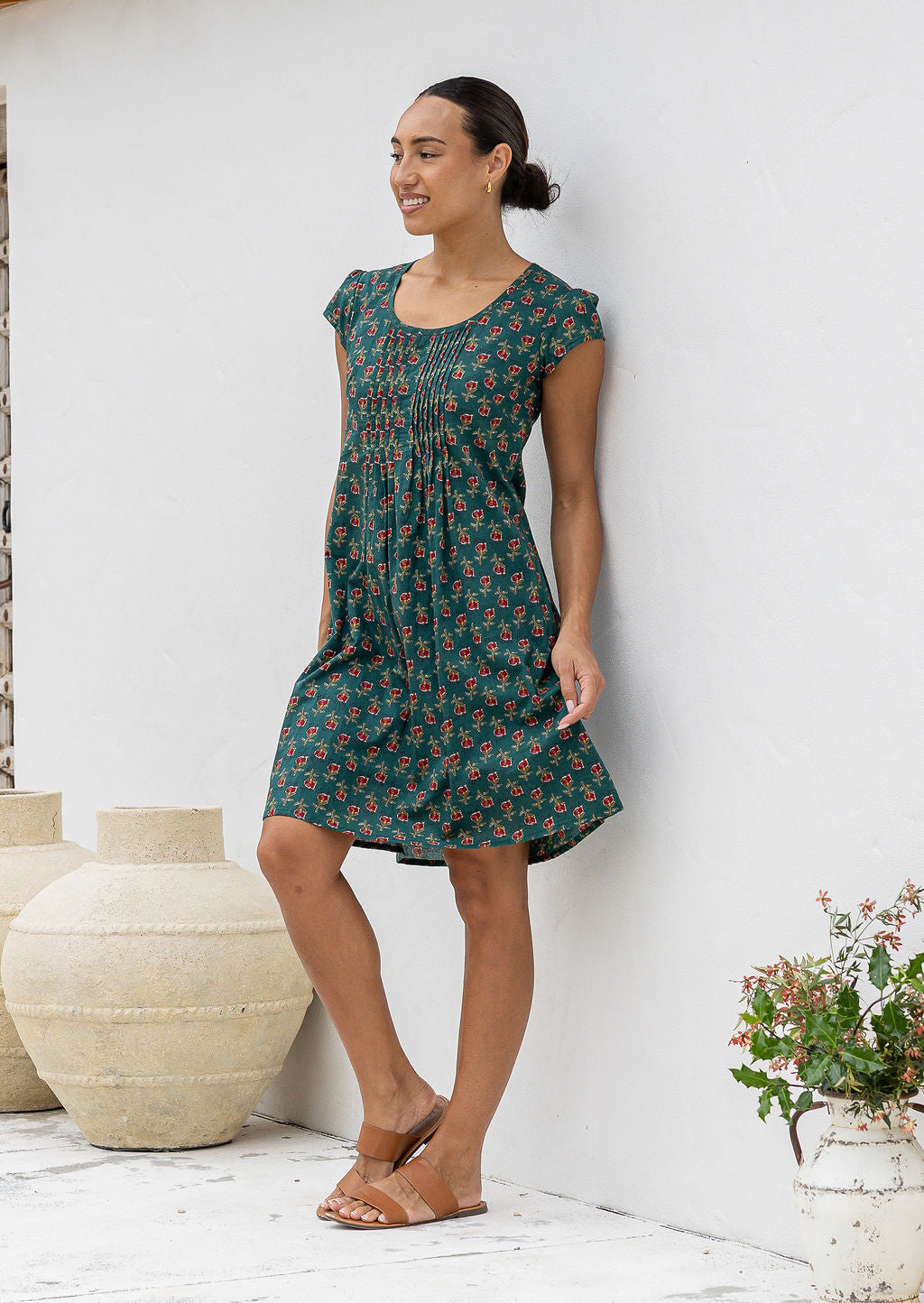 Woman wearing a dark green cotton floral dress standing against a white wall with decorative vases and plants.