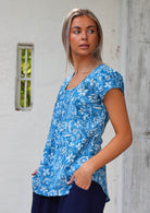 Woman stands sideways in cotton cap sleeved top with scoop round neckline, paired with dark blue cotton pants and her hands in the pockets