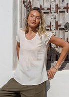 Woman wearing a white cotton and olive green pants standing in front of a rustic wooden door.