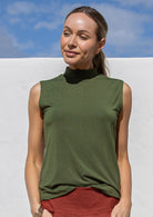 Woman wearing an olive green sleeveless top with a high neckline against a blue sky and white wall.