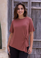 Woman wearing a terracotta short sleeved rayon top with a knot detail, standing against a wooden door