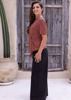 Woman stands sideways wearing a terracotta short sleeve rayon top with a knot detail in the front paired with black pants