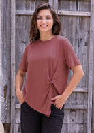 Woman wearing a terracotta rayon top with a knot detail in the front, standing against a wooden wall.