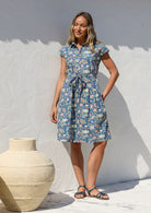 Model stands with one hand in pocket wearing a 100% cotton knee length shirt style retro floral dress in dusty blue