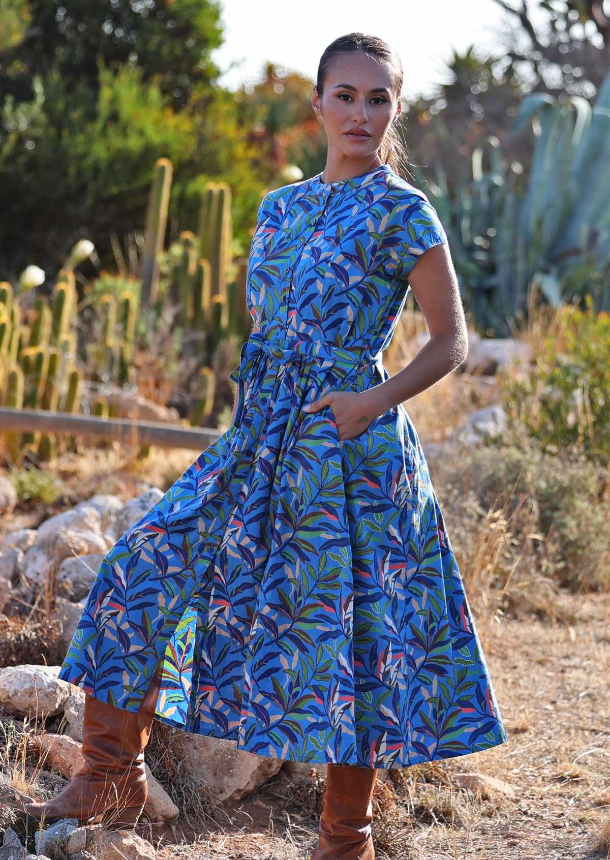 Woman standing with one foot on a rock in blue cotton leafy button down maxi dress with her hands in pockets 