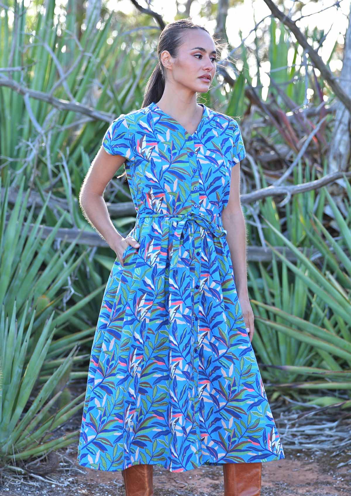 Woman standing facing to the side in blue cotton leafy button down maxi dress with one hand in pocket 