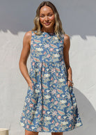 Woman in a blue floral cotton dress with sleeveless relaxed fit design and hands in pockets, standing next to a large vase against a white wall