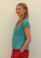 Model smiling with one hand on in pant pocket standing to the side wearing 100 percent cotton top in aqua with small functional shell buttons