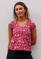 Woman wears cotton cap sleeve top with scoop neckline and decorative pleats across chest