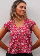 Woman leans on wall in cotton cap sleeve floral print top with decorative pin tucks across chest and coconut buttons