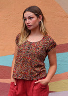 Model facing to the side hands in pant pockets wearing 100 percent cotton Indian block print blouse florals on a red base with buttons