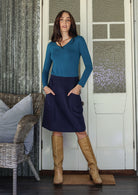 Woman wearing a teal top and navy cotton corduroy skirt with tan boots standing with hands in pockets