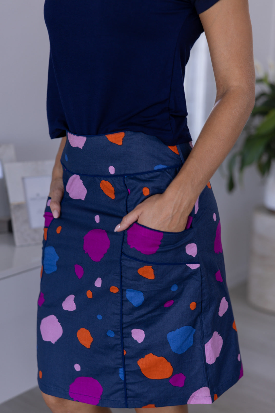 Close up picture of a woman standing in a modern room, wearing a knee-length skirt with a multicoloured spot print, with her hands in pocket