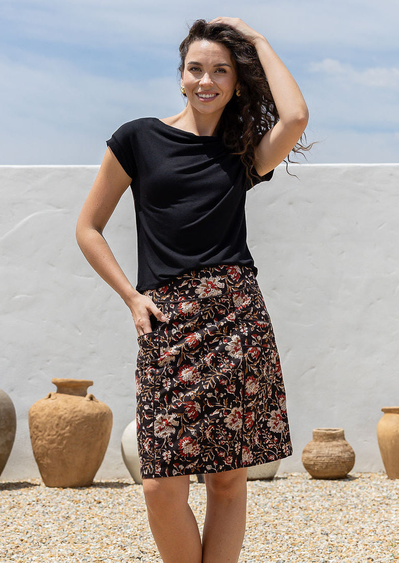 Woman in a black top and cotton dark  floral skirt standing outdoors with pottery in the background