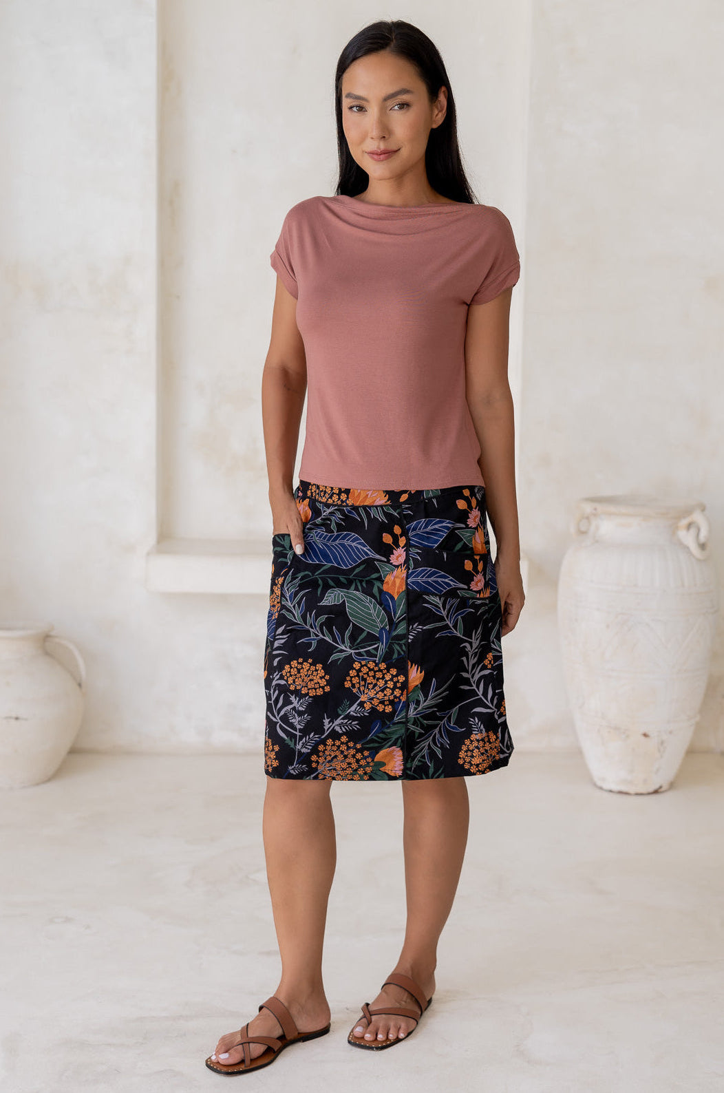Woman standing with hand in pocket wears black cotton above knee length skirt with colorful botanical print and piped detailing.