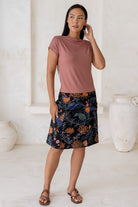 Woman wearing a dusty pink top and floral  cotton skirt in black with A-line shape, piped detailing and pockets.