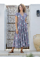 Woman poses in blue based floral print rayon maxi dress with V-neckline, buttoned bodice, waist-tie and pockets