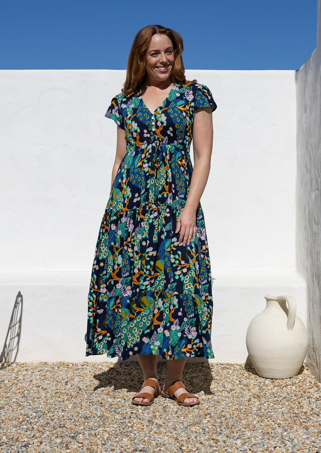 A woman wearing a short-sleeve, V-neck rayon maxi dress with a colourful floral peacock print on a dark navy background, standing outdoors against a white wall.