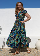 A woman wearing a short-sleeve, V-neck maxi dress with a buttoned bodice and shoestring ties to cinch the waist, with hands in pockets