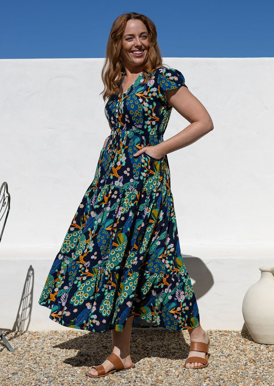 A woman wearing a short-sleeve, V-neck maxi dress with a buttoned bodice and shoestring ties to cinch the waist, with hands in pockets