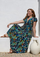 Woman wearing a short-sleeve maxi dress with a colourful peacock print in teal, blue, yellow, and orange, sitting on a white stone ledge next to a white ceramic jug.