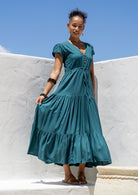 Woman in a teal maxi dress with full skirt standing against a white wall with blue sky.