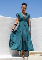 Woman in a teal maxi dress with v-neck standing against a white wall with blue sky.