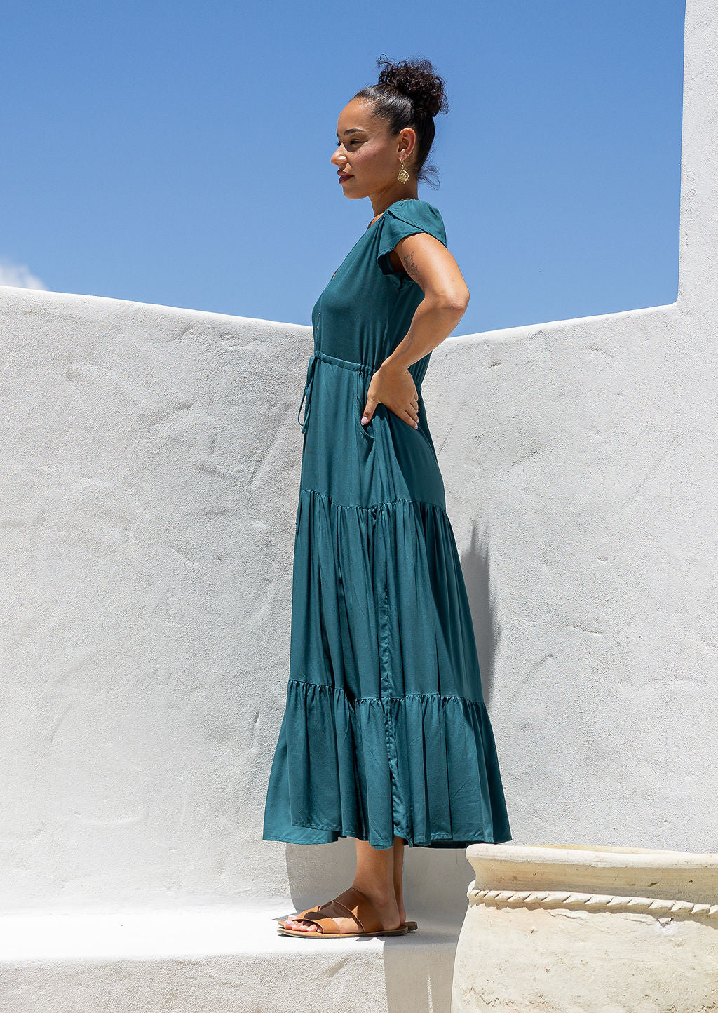 Woman in a teal maxi dress standing against a white wall with blue sky with hand on hip