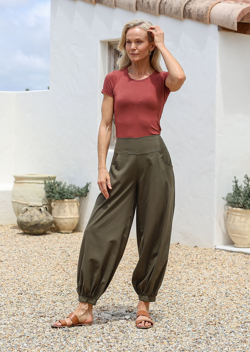 Woman standing outdoors wears 100% cotton relaxed fit pants in olive green with pockets, elastic at back of waist and cuffed ankles.