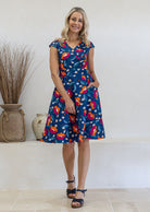 Model with one hand in pocket wears 100% cotton dress with crossover bodice, cap sleeves and fuller skirt in royal blue based floral print.