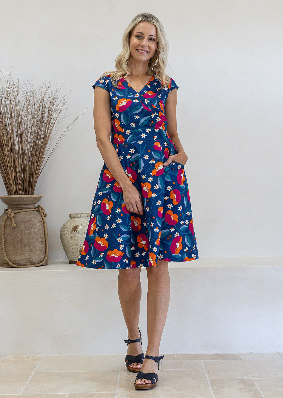 Model with one hand in pocket wears 100% cotton dress with crossover bodice, cap sleeves and fuller skirt in royal blue based floral print.