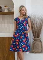 A woman wearing a blue wrap-style dress with a colorful floral print, standing in a bright indoor setting next to a vase of dried grass.