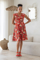 Woman wearing a cotton above knee dress with cap sleeves, pockets and generous skirt, poses in a living room setting.