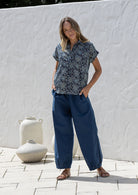 Woman wearing a navy blue based floral 100% cotton top and plain pants standing in a rustic indoor setting.