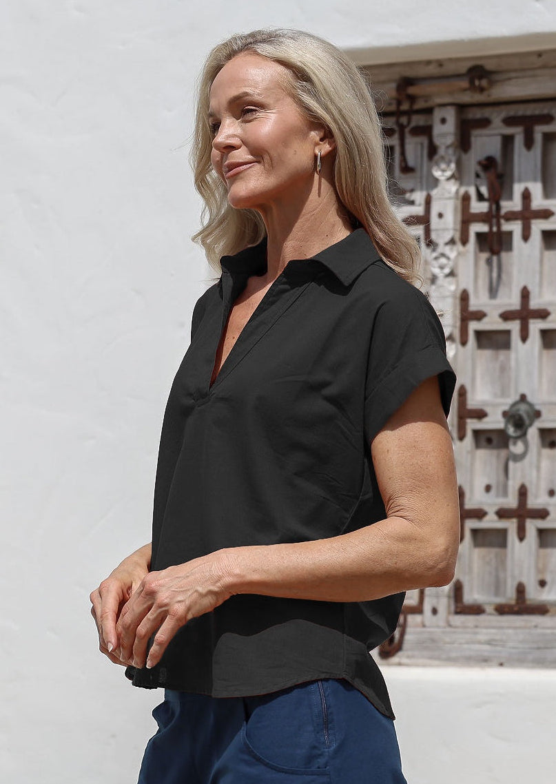Woman wearing a black cotton collared short sleeved shirt and blue pants standing in front of a white wall