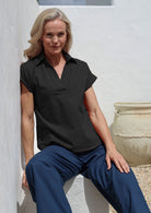 Woman wearing a black cotton short sleeved collared top and blue pants sitting against a white wall.