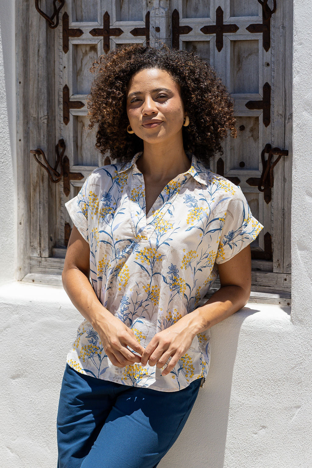 Woman wearing a neutral based floral 100% cotton top and blue pants in front of a wooden door.