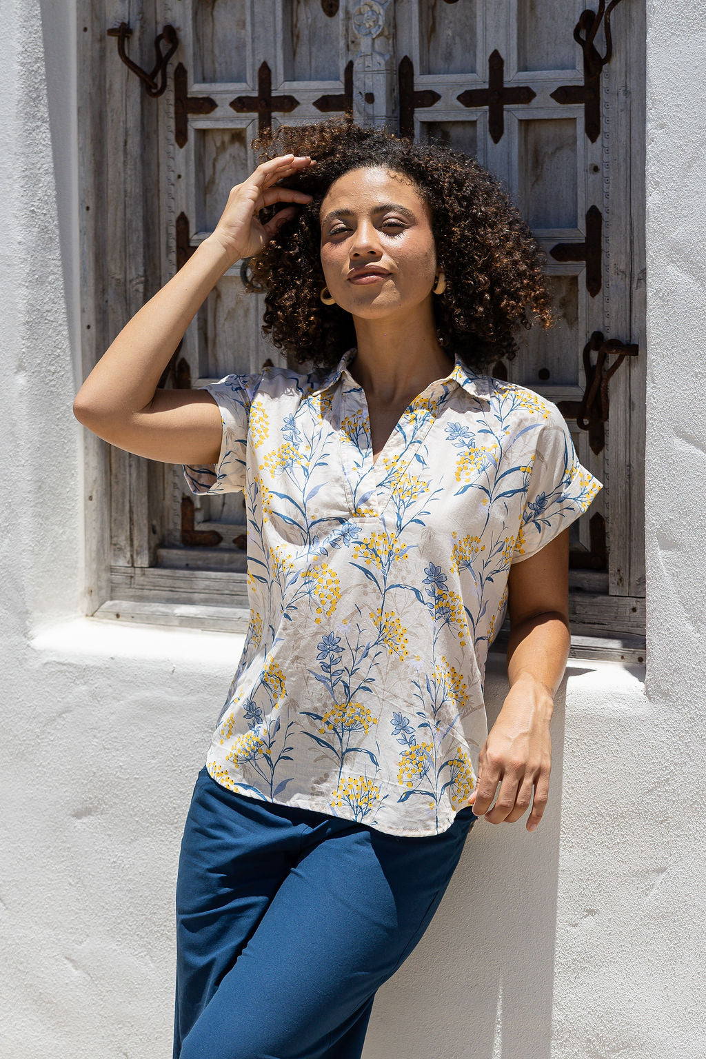 Woman stands in front of a wooden door wearing a neutral based floral cotton top with short folded cuff sleeves, collar and V neckline 